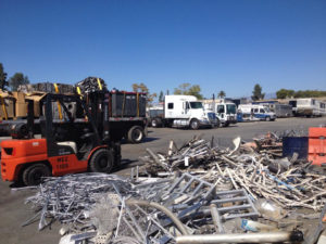 A busy recycling yard with piles of scrap metal, a forklift, and trucks at MEC Recycling LLC in Bakersfield, CA.