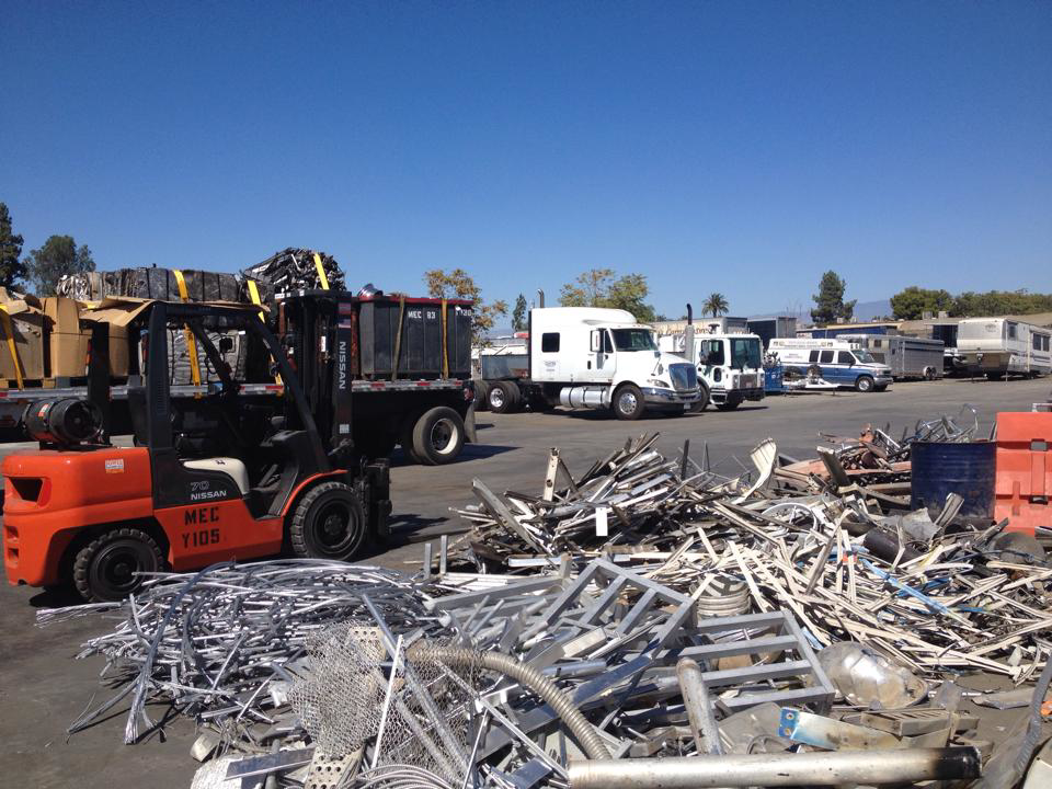 A busy recycling yard with piles of scrap metal, a forklift, and trucks at MEC Recycling LLC in Bakersfield, CA.